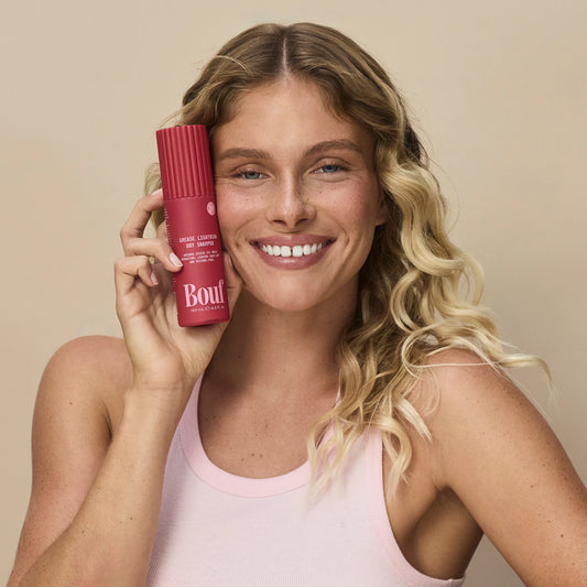 A smiling blonde woman holds a pink cosmetic bottle next to her face against a plain beige background. The bottle has a ribbed texture and features the brand name 'Bouf' in white.