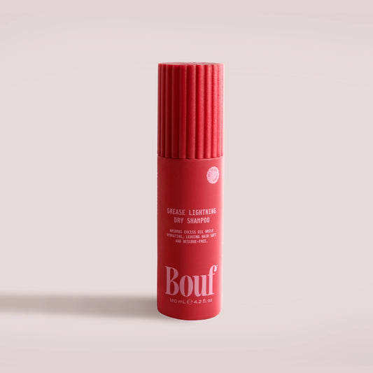 A red cylindrical bottle of Bouf Grease Lightening Dry Shampoo, featuring a ribbed cap and white text, set against a solid pale pink background.