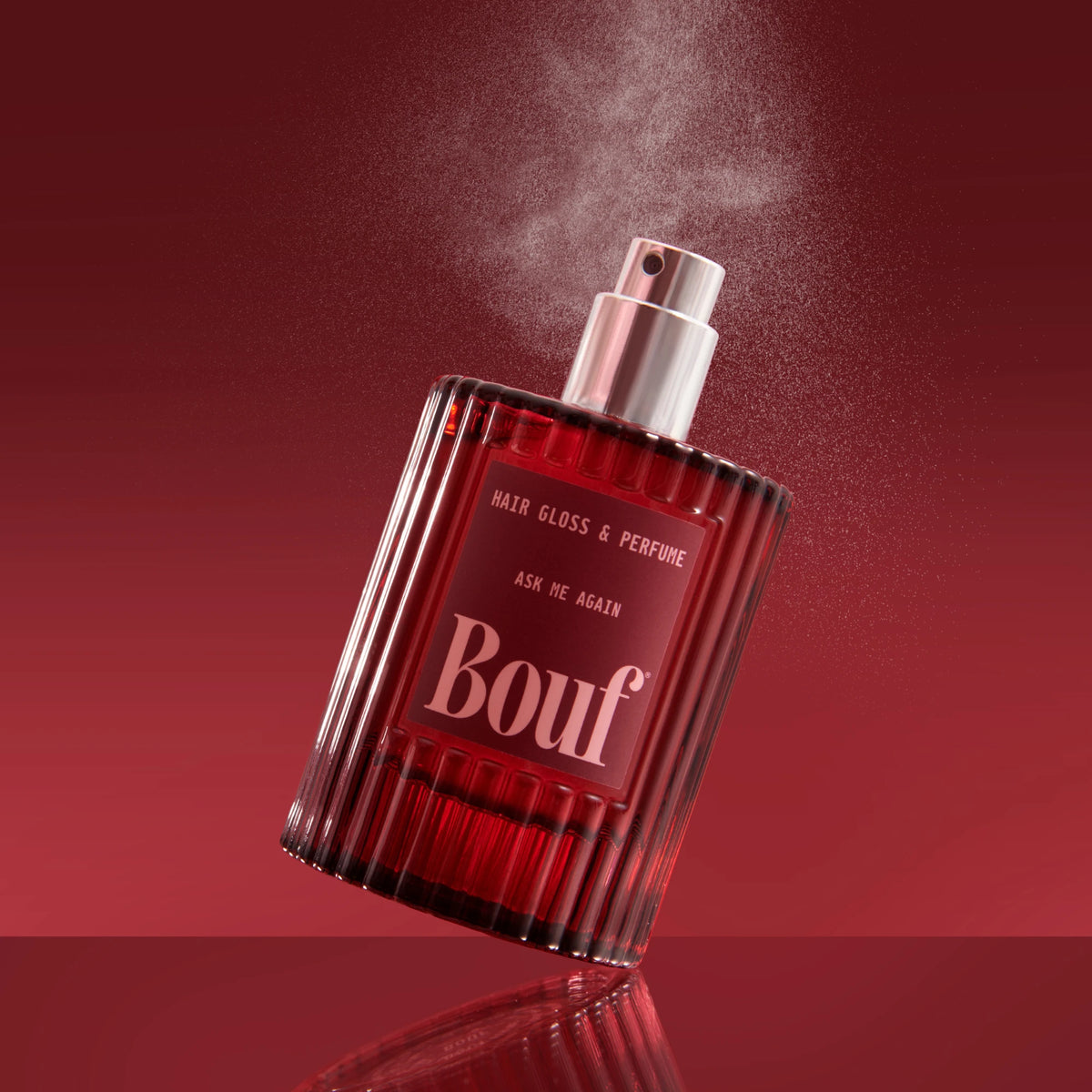 A bottle of Bouf hair gloss and perfume with a metallic silver spray nozzle, shown spraying with fine mist against a dark red gradient background.