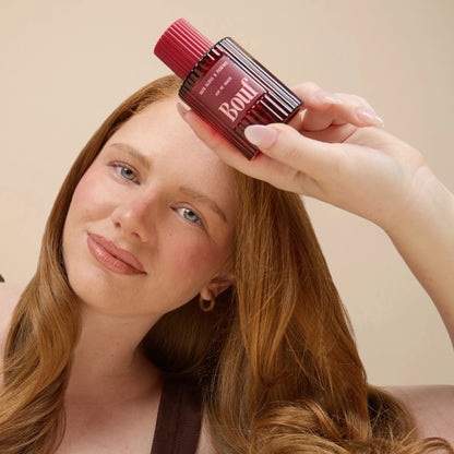 A woman with long red hair holding a bottle of Bouf Hair Gloss & Perfume against a plain beige background.