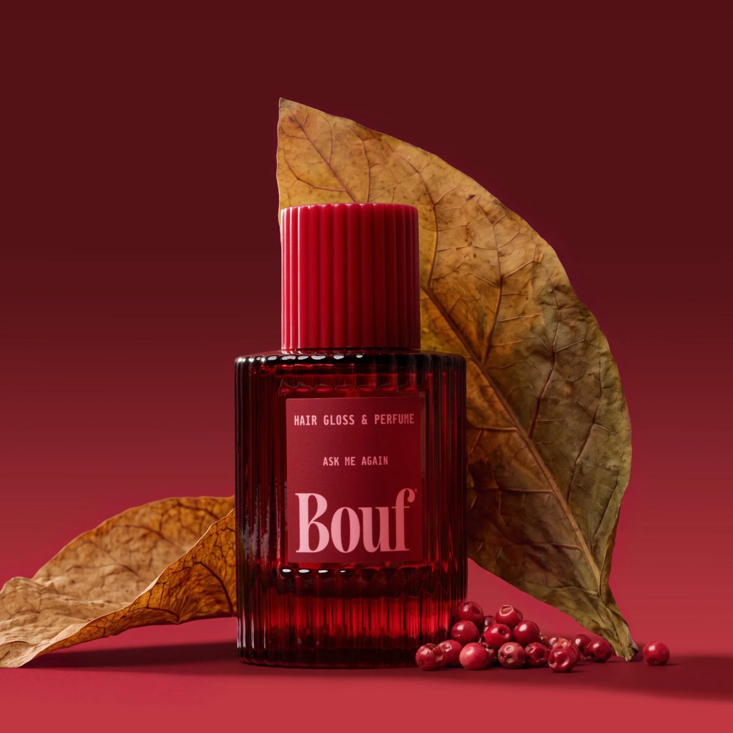 Bouf hair gloss and perfume bottle styled with tabacco leaves and small red berries on a dark red background.