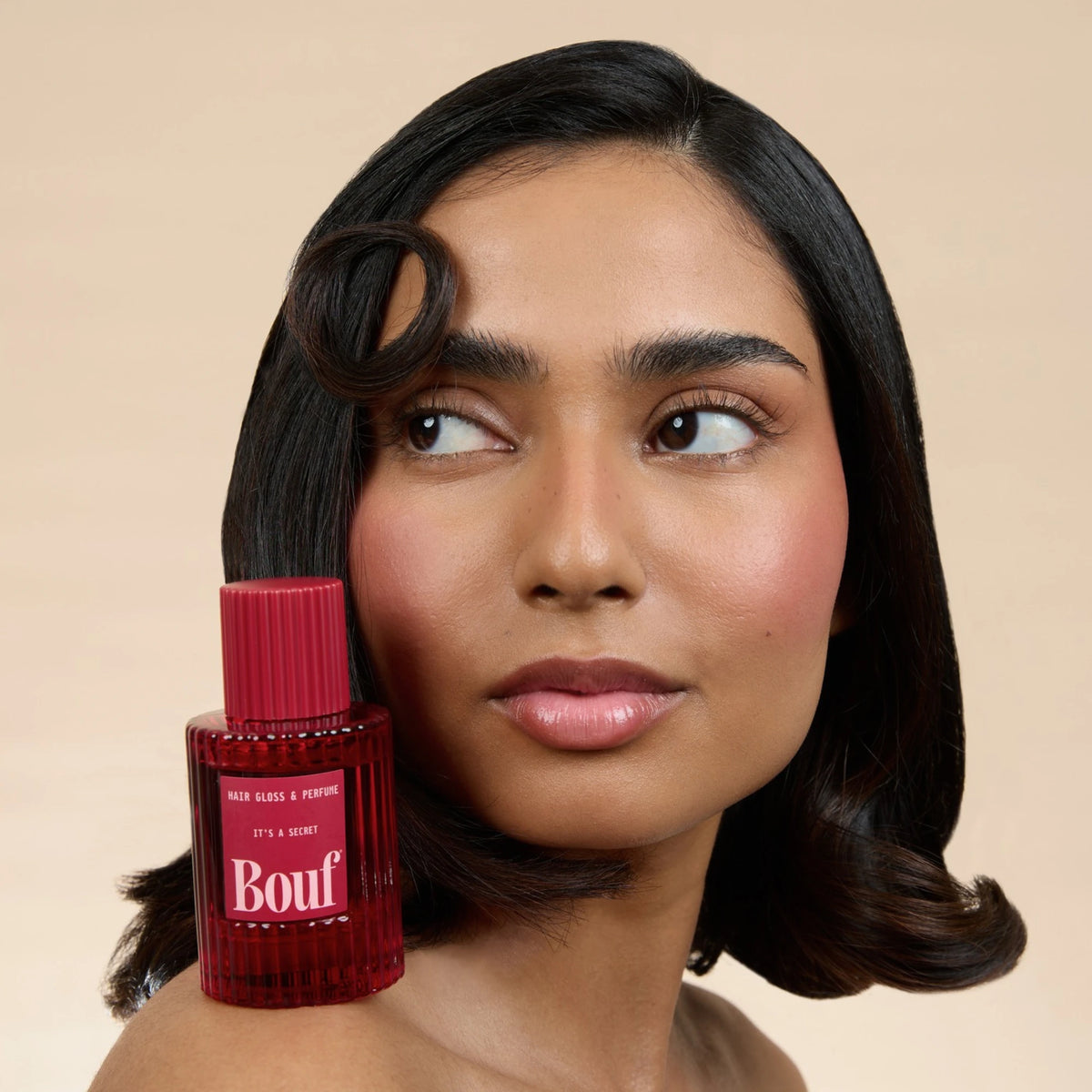 A woman with styled dark hair with a bottle of Bouf Hair Gloss & Perfume on her shoulder against a solid beige background.