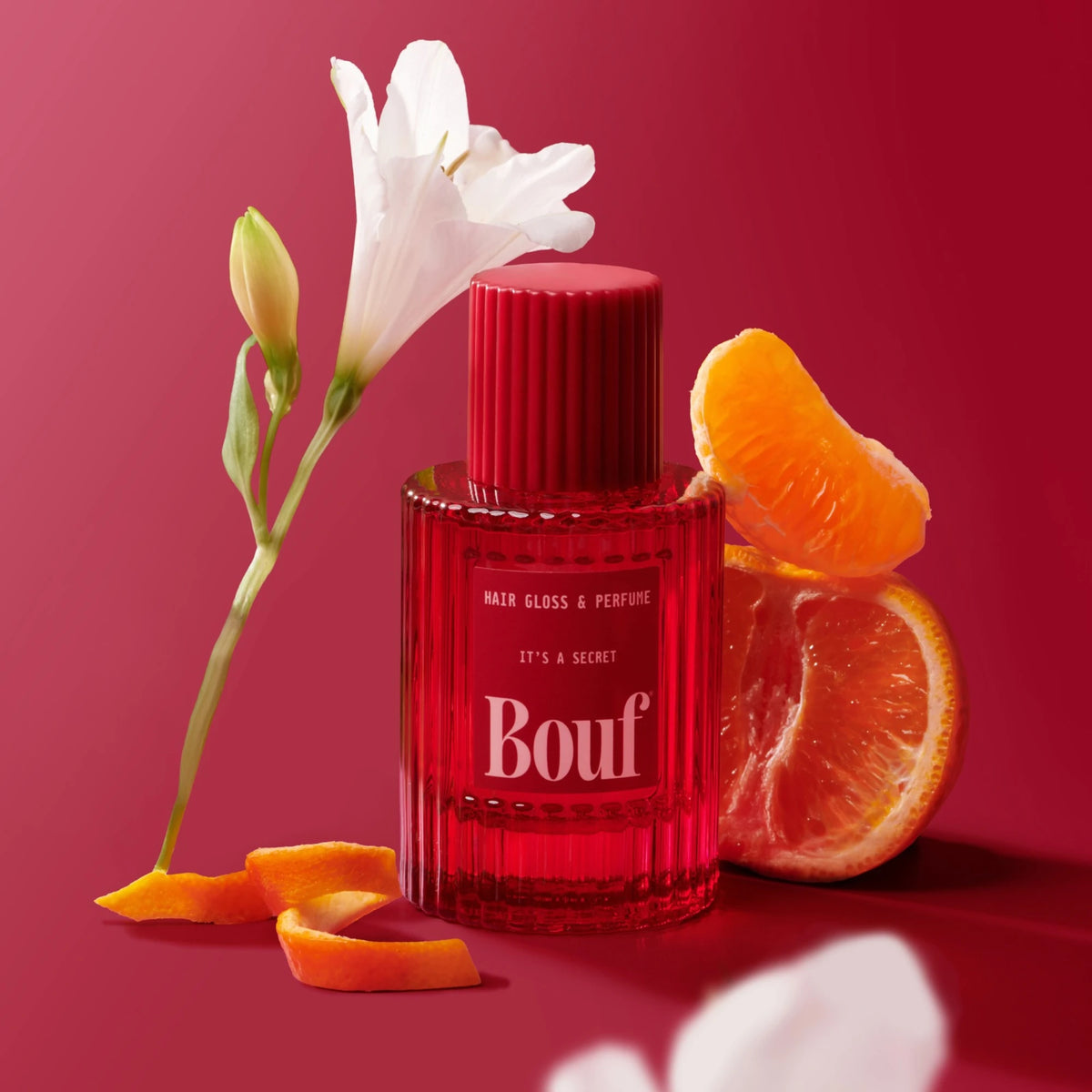 A bottle of Bouf hair gloss and perfume with a white fresha and mandarin segments on a deep pink background.