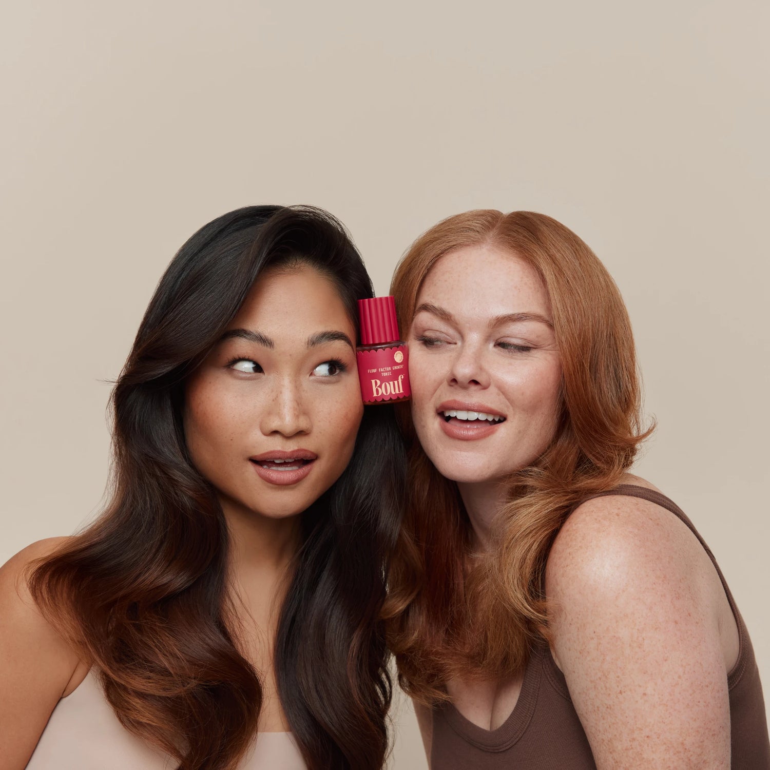 Two women posing closely together against a solid beige background, with a small pink bottle of Bouf hair product tucked between their hair.