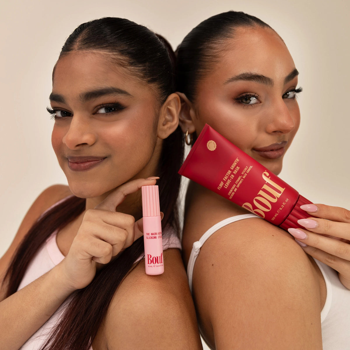 Two women posing back-to-back, each holding a product from Bouf's Slick Me Baby Bundle — the Hairmaster Slicking Stick and the Flouf Factor Growth Leave-In Mask — against a warm beige background