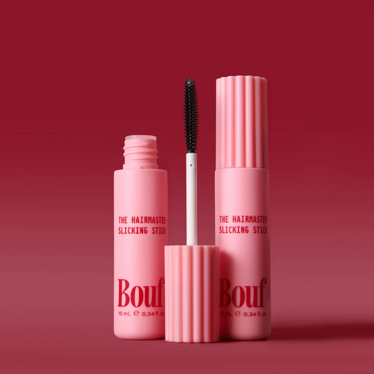 Bouf The Hairmaster Slicking Stick in a matte pink bottle with a fluted cap, shown alongside the applicator wand and a matching cap on a dark red gradient background.